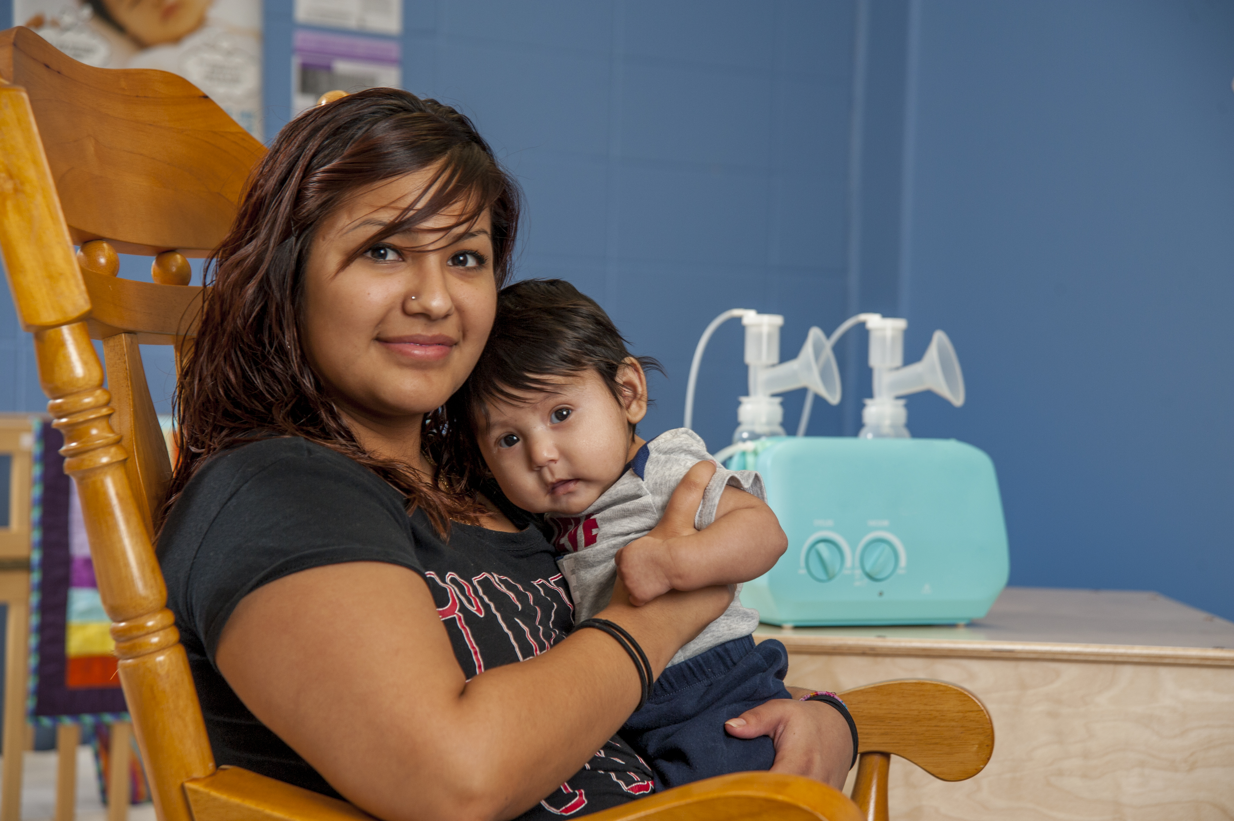A smiling mother and baby sit in a rocking chair. A breast pump sits on a table in the background.
