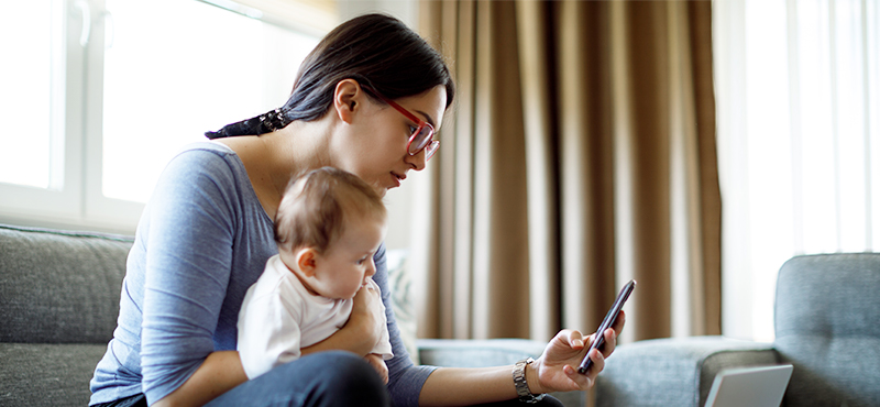 Mother sitting and holding her infant while looking at her phone and a laptop.